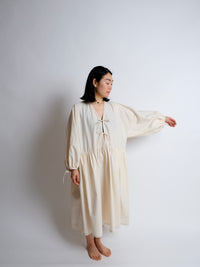 Woman wearing the Bow Dress Hack sewing pattern from Birgitta Helmersson on The Fold Line. An oversized dress pattern made in cotton or linen fabric, featuring a V-neckline, tie front, voluminous long sleeves with ties, a gathered skirt with pockets, and midi length.