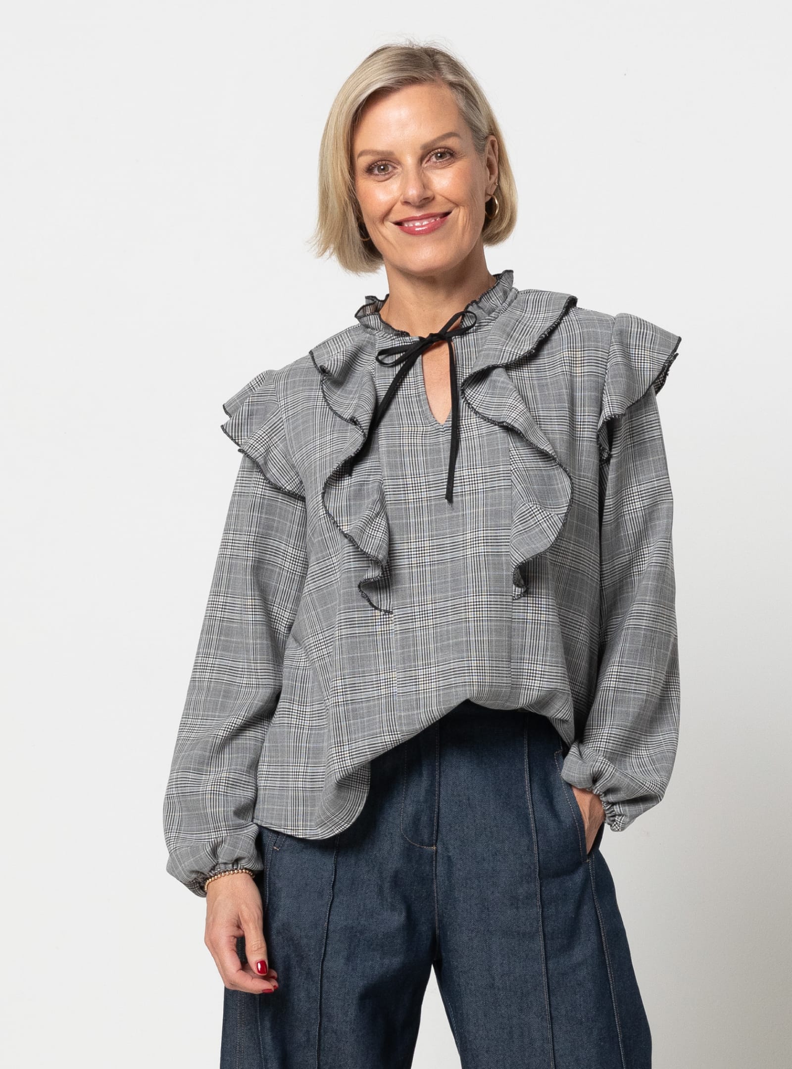 Woman wearing the Cendrine Woven Blouse Set sewing pattern from Style Arc on The Fold Line. A top pattern made in seersucker, chambray, cotton lawn, crepe, or georgette fabric, featuring a ruffled round neckline with a front split and tie, bodice and shoulder frills, and long sleeves.