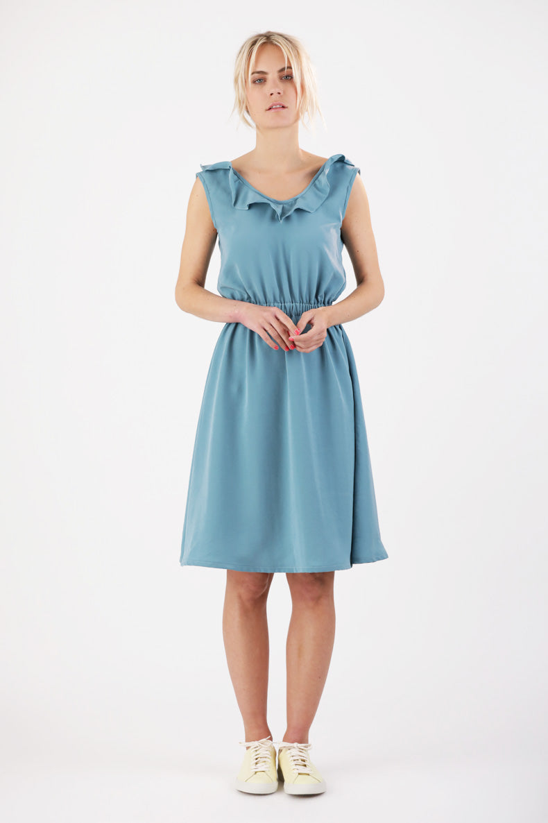 Woman wearing the Chari Dress sewing pattern from Schnittchen Patterns on The Fold Line. A sleeveless dress pattern made in light cotton, viscose, linen, or light denim fabric, featuring a ruffled V-neckline, elasticated waist, and knee length.