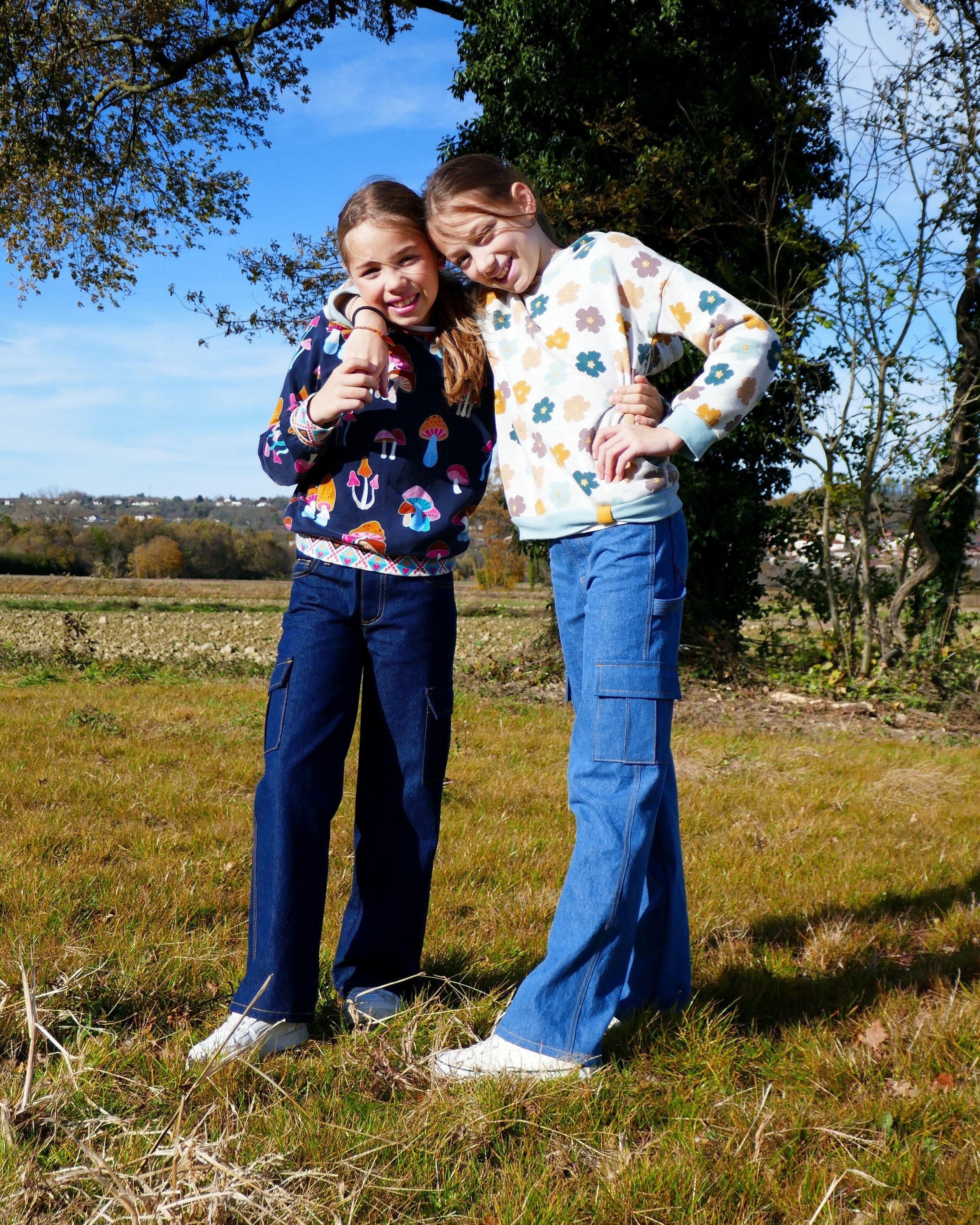 Girls wearing the Django Jeans or Cargo Pants sewing pattern from Petits D'om on The Fold Line. A pants pattern made in denim or twill fabric, featuring a zipper fly and buttoned waistband, front pockets with a coin pocket, back patch pockets with a hammer loop, side (cargo) pockets, belt loops, and rivets.