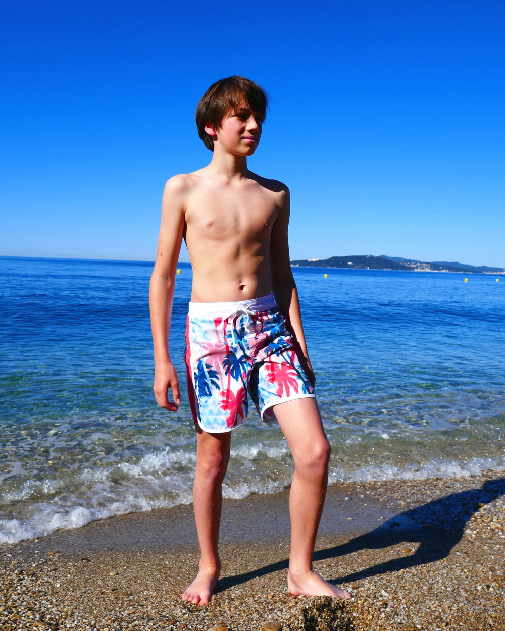 Boy wearing the Child/Teen Huanchaco Swim Shorts sewing pattern from Petits D'om on The Fold Line. A swim shorts pattern made in polyester fabric, featuring a faux fly, flat front waistband with a drawstring, elasticated back waistband, back yoke, side panels, bias bound edges, an inner mesh brief, and mid-thigh length.