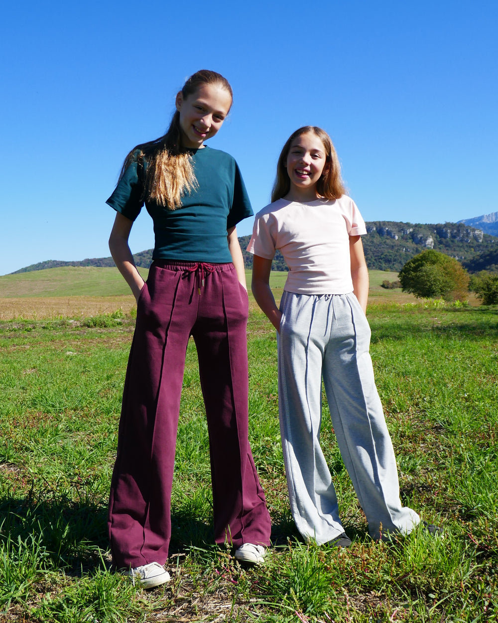 Girls wearing the Child/Teen Norn Sweatpants sewing pattern from Petits D'om on The Fold Line. A sweatpants pattern made in fleece or french terry fabric, featuring an elastic waist with a drawstring, pockets, and wide legs with a vertical pintuck down the centre front of each leg.