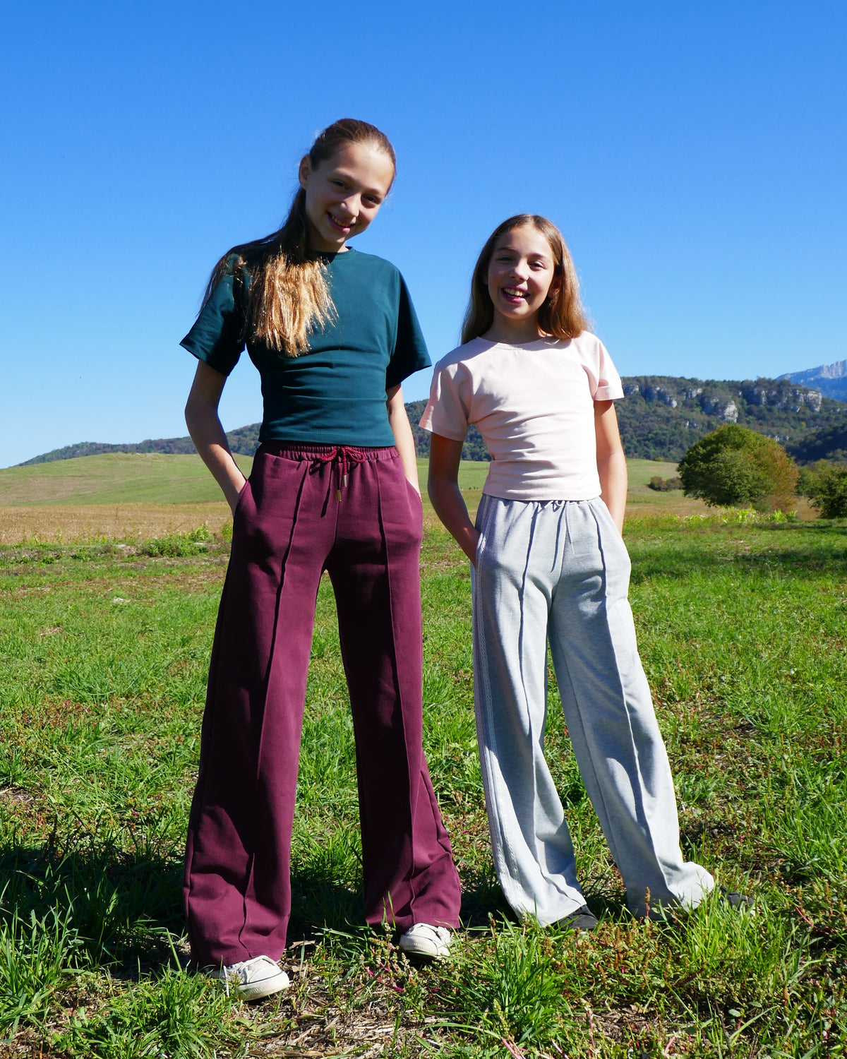 Girls wearing the Child/Teen Norn Sweatpants sewing pattern from Petits D'om on The Fold Line. A sweatpants pattern made in fleece or french terry fabric, featuring an elastic waist with a drawstring, pockets, and wide legs with a vertical pintuck down the centre front of each leg.