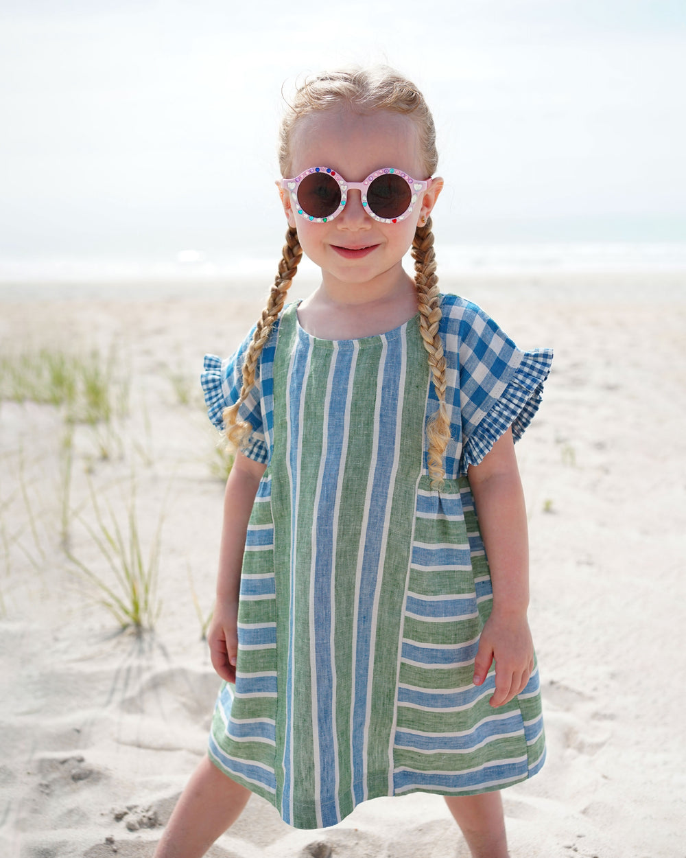 Girl wearing the Children's Collage Gather Dress sewing pattern from Matchy Matchy Sewing Club on The Fold Line. A dress pattern made in cotton, linen, twill, rayon, or viscose fabric, featuring a relaxed fit, round neckline, short sleeves with ruffles, side gathers, and back button placket.