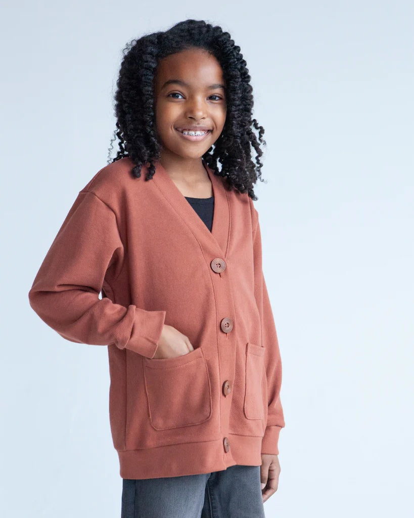 Girl wearing the Children's Mini Marlo Sweater sewing pattern from True Bias on The Fold Line. A cardigan pattern made in sweater knit, french terry, sweatshirting, polar fleece, rib knit, or boucle fabric, featuring an oversized fit, dropped shoulder, deep V-neck, wide bands, large buttons, and patch pockets.