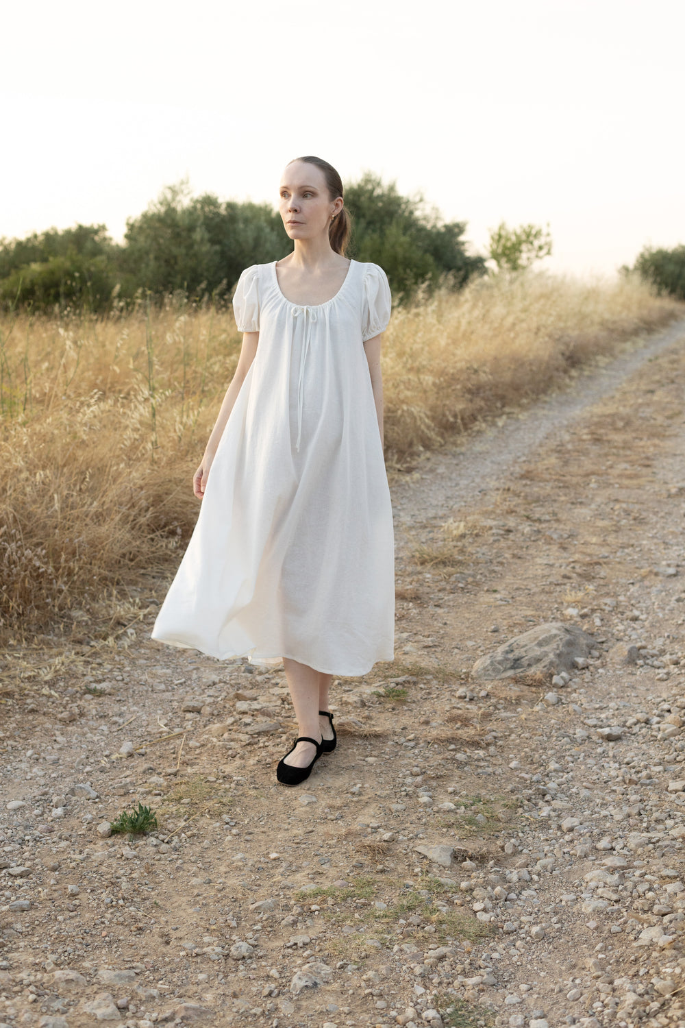 Woman wearing the Chloe Dress sewing pattern from Silversaga Patterns on The Fold Line. A vintage-inspired dress pattern made in cotton or linen fabric, featuring an adjustable scoop neckline with fabric ties, short puff sleeves, hidden in-seam pockets, and midi length.