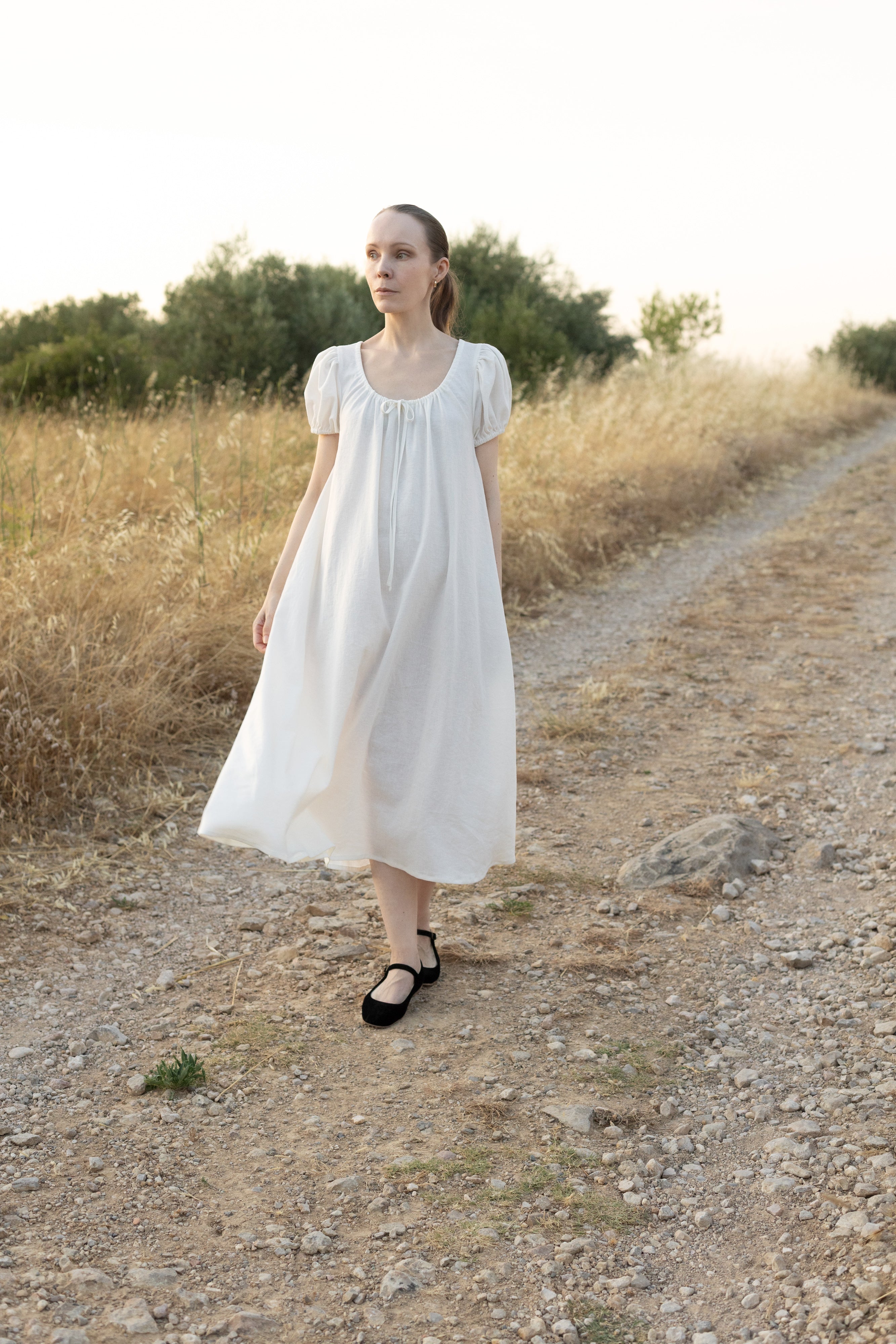 Woman wearing the Chloe Dress sewing pattern from Silversaga Patterns on The Fold Line. A vintage-inspired dress pattern made in cotton or linen fabric, featuring an adjustable scoop neckline with fabric ties, short puff sleeves, hidden in-seam pockets, and midi length.