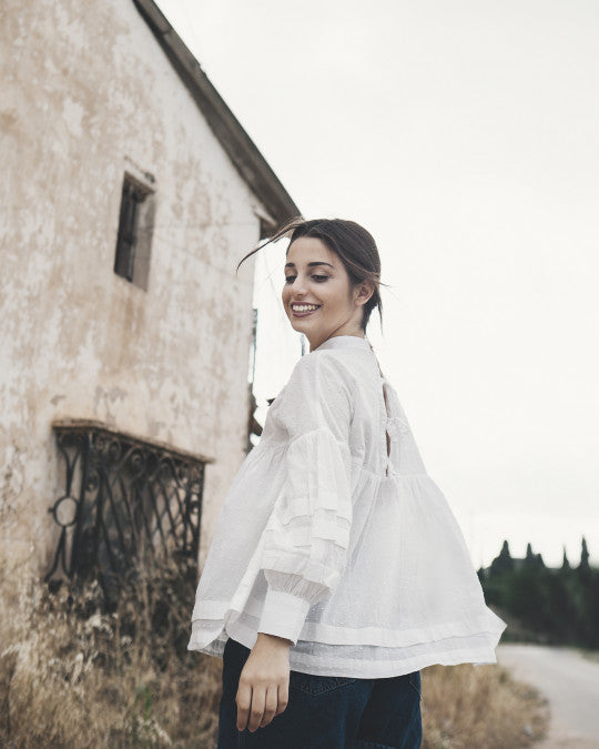 Woman wearing the Coeli Blouse sewing pattern from Pauline Alice on The Fold Line. A blouse pattern made in cotton lawn, silk, swiss dot, viscose, or voile fabric, featuring a loose smock silhouette, high collar, shoulder yoke, wide horizontal knife pleats, billowing sleeves gathered into cuffs, and self ties in the back.
