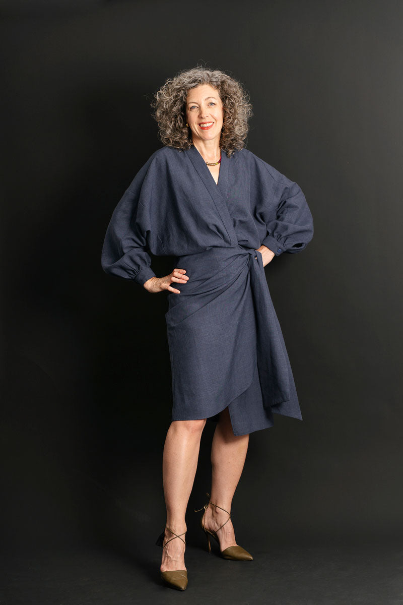 Woman wearing the Copenhagen Wrap Dress sewing pattern from Liesl + Co on The Fold Line. A wrap dress pattern made in cotton poplin, lightweight twill, shirtings, sateen, linen, or double gauze fabric, featuring a blousey top, V-neckline, long voluminous sleeves with cuffs, sarong-style skirt, and knee length.