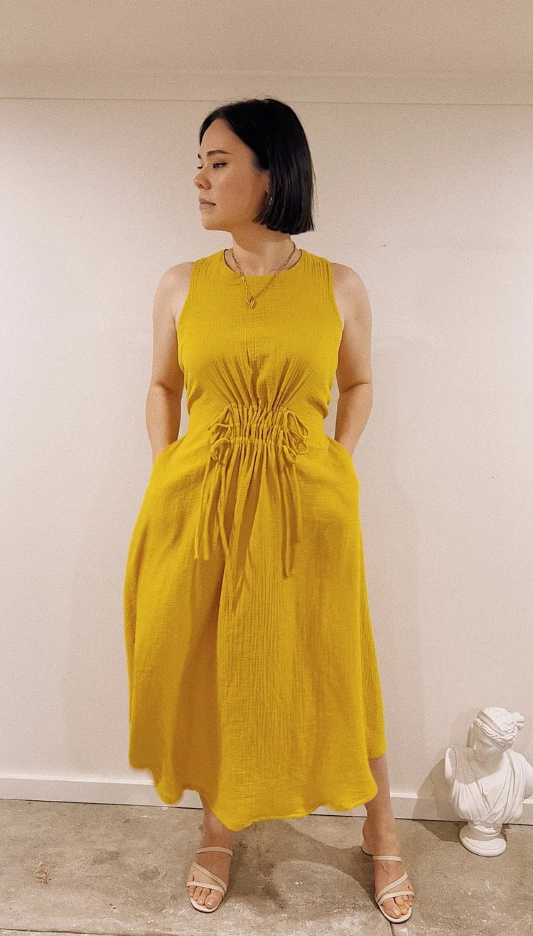 Woman wearing the Cordelia Dress sewing pattern from Veronica Tucker on The Fold Line. A sleeveless dress pattern made in linen or cotton fabric, featuring a round neckline, keyhole back opening, drawstring waist detail, pockets, and midi length.