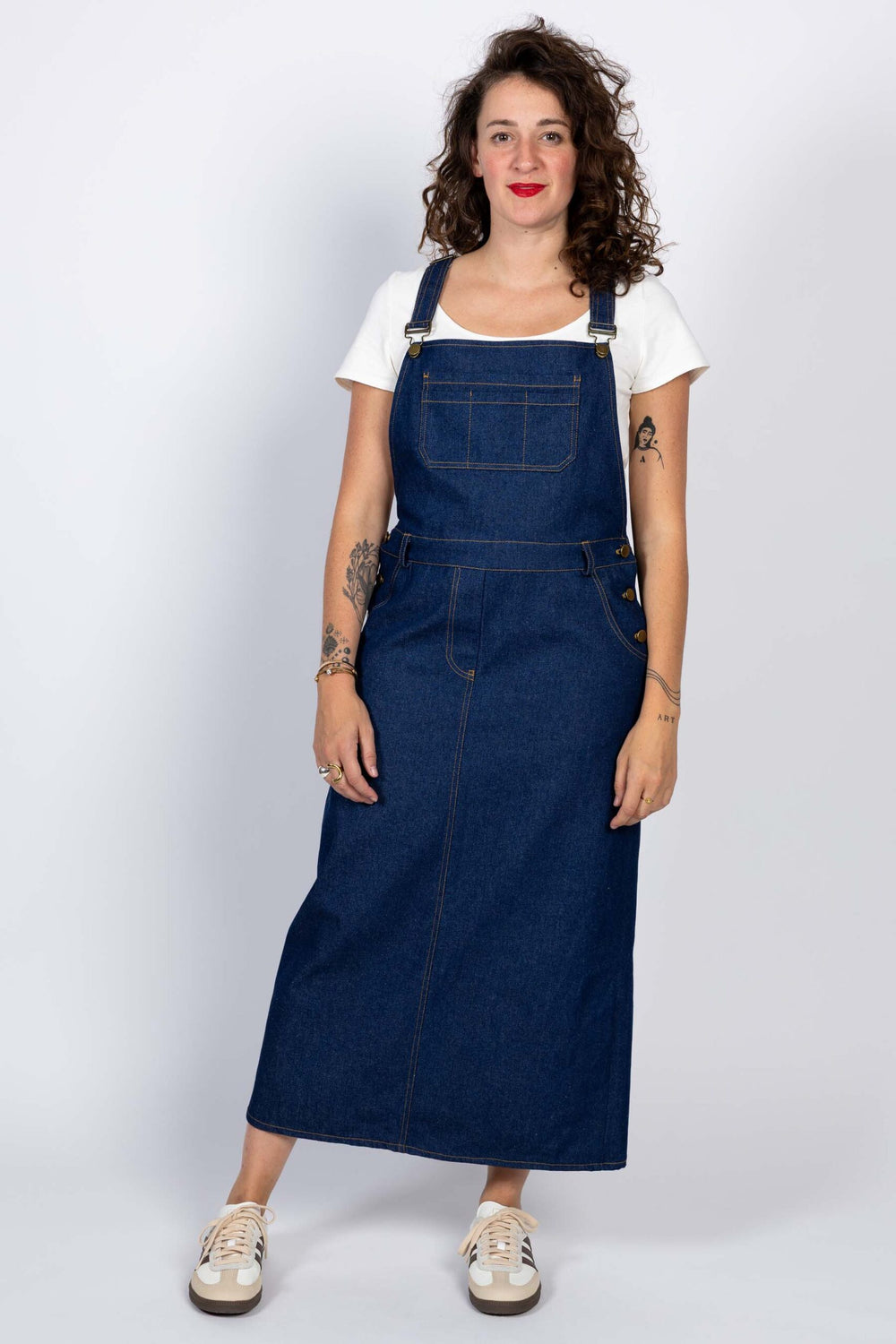 Woman wearing the Crystal Dungaree Dress sewing pattern from I AM Patterns on The Fold Line. A dungaree dress pattern made in denim, cotton canvas, gabardine, corduroy, or cotton twill fabric, featuring a loose fit, front and back bib, adjustable straps, chest pocket, front slash pockets, buttoned side openings, faux fly, contrast topstitching, and maxi length.