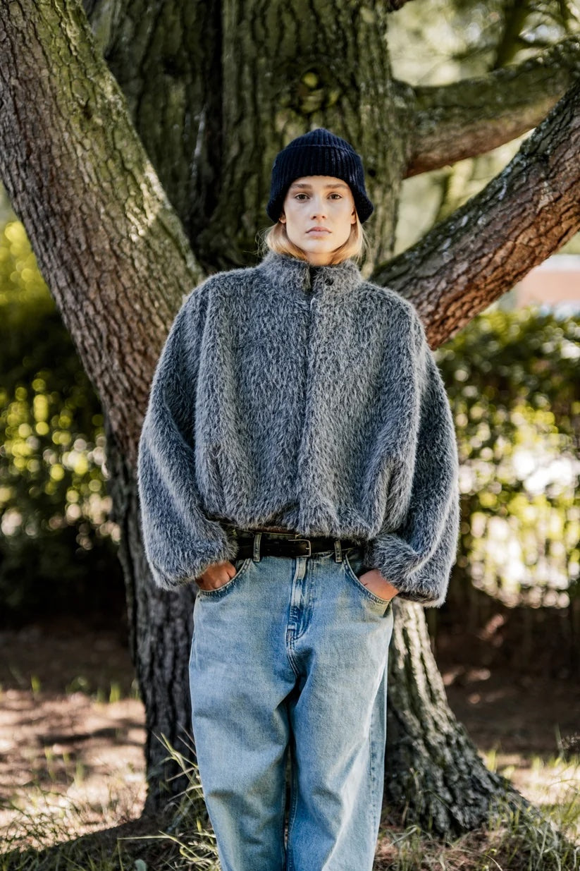 Woman wearing the Dante Jacket sewing pattern from Fibre Mood on The Fold Line. A bomber jacket pattern made in shaggy (faux) fur, teddy, bouclé, or fur-effect fabric, featuring a rounded silhouette, high neck, concealed button placket, pockets, and voluminous sleeves with elasticated cuffs.