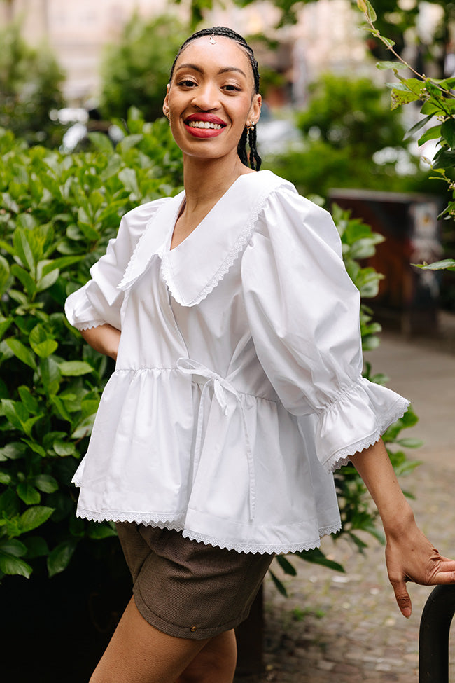 Woman wearing the Daphne Wrap Blouse sewing pattern from JULIANA MARTEJEVS on The Fold Line. A top pattern made in cotton fabric, featuring an oversized silhouette, V-neckline, statement collar, wrap front with a tie, gathered peplum, mid-length puff sleeves, and scalloped trim.