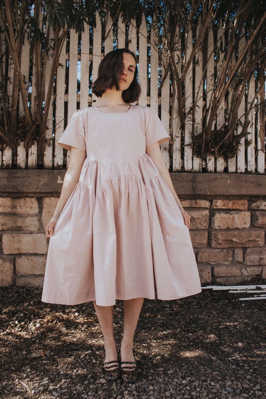 Woman wearing the Delft Dress sewing pattern from Madswick on The Fold Line. A dress pattern made in linen, chambray, cotton lawn, cotton poplin, seersucker, or cotton twill fabric, featuring a rounded square neckline, short puff sleeves, a curved waistline, scalloped gathered skirt panels, and knee length.