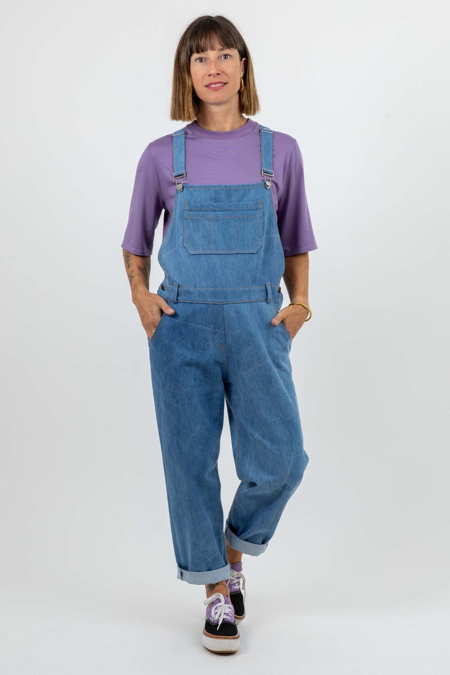 Woman wearing the Diamond Dungarees sewing pattern from I AM Patterns on The Fold Line. An overalls pattern made in denim, cotton canvas, gabardine, corduroy, or cotton twill fabric, featuring a loose fit, front and back bib, adjustable straps, chest pocket, front slash pockets, back patch pockets, buttoned side openings, faux fly, and contrast topstitching.