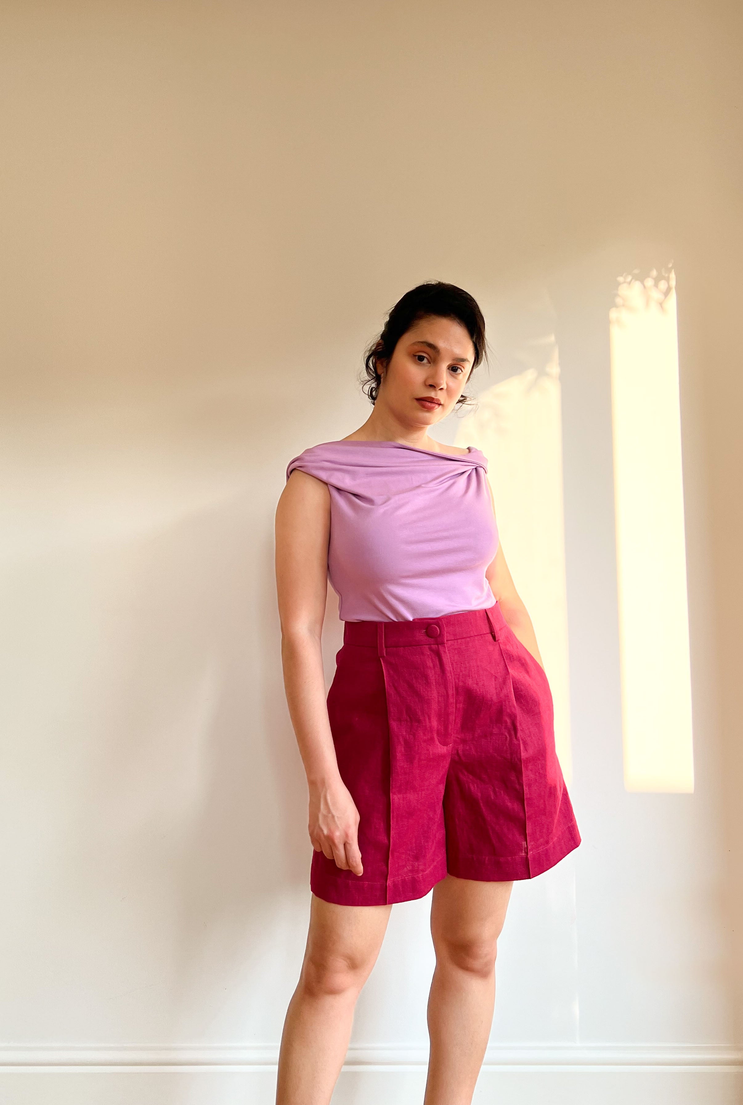 Woman wearing the Donna Bermuda Shorts sewing pattern from Bella Loves Patterns on The Fold Line. A shorts pattern made in linen or suiting fabric, featuring a flat waistband at the front, elastic at the back, fly front zip, front pleats, topstitched crease lines, in-seam side pockets, and a wide topstitched hem.