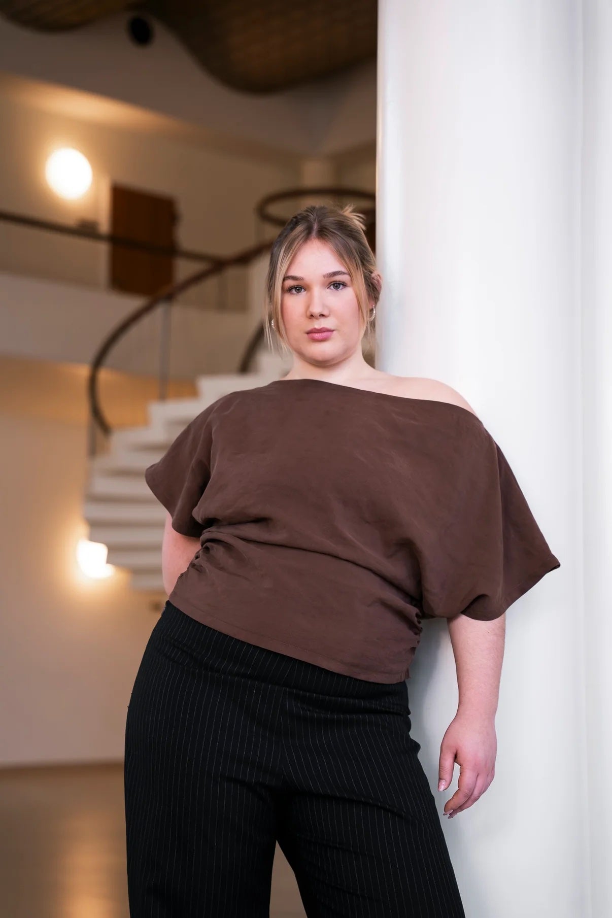 Woman wearing the Drop Top sewing pattern from Puff and Pencil on The Fold Line. A top pattern made in linen, poplin, woven viscose, thin wool, chiffon, crepe, organza, taslan, or satin fabric, featuring a one shoulder silhouette, wide neckline, short grown-on sleeves, and ruched side seams.