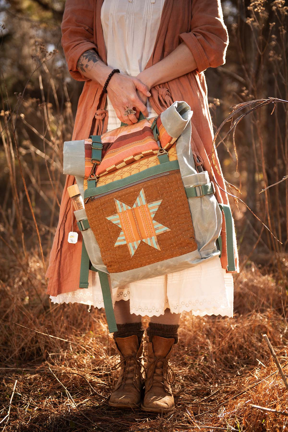 Woman holding the Elevenses Backpack sewing pattern from Sew Liberated on The Fold Line. A backpack pattern made in canvas, oiled canvas, waxed canvas, or twill fabric, featuring a quilted panel, rolled top, side water bottle pockets, a front zipper pocket, and padded straps.