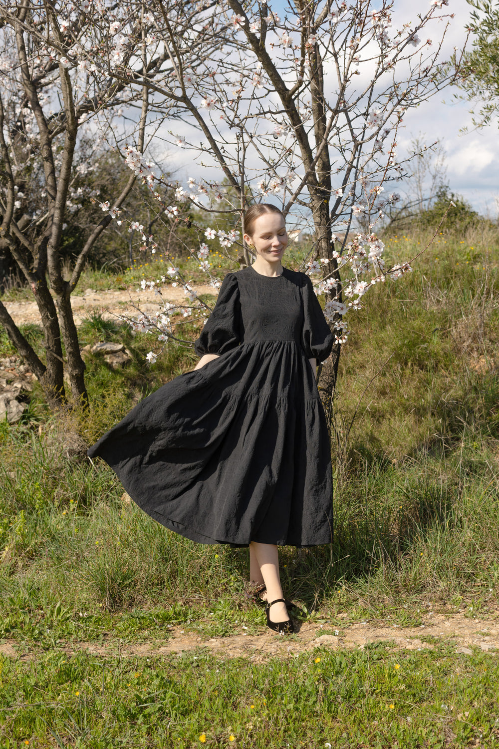 Woman wearing the Elsa Dress sewing pattern from Silversaga Patterns on The Fold Line. A dress pattern made in cotton, linen, or wool fabric, featuring a round neckline, mid-length statement sleeves with a dipped hemline, high curved waistline, and a tiered midi length gathered skirt.