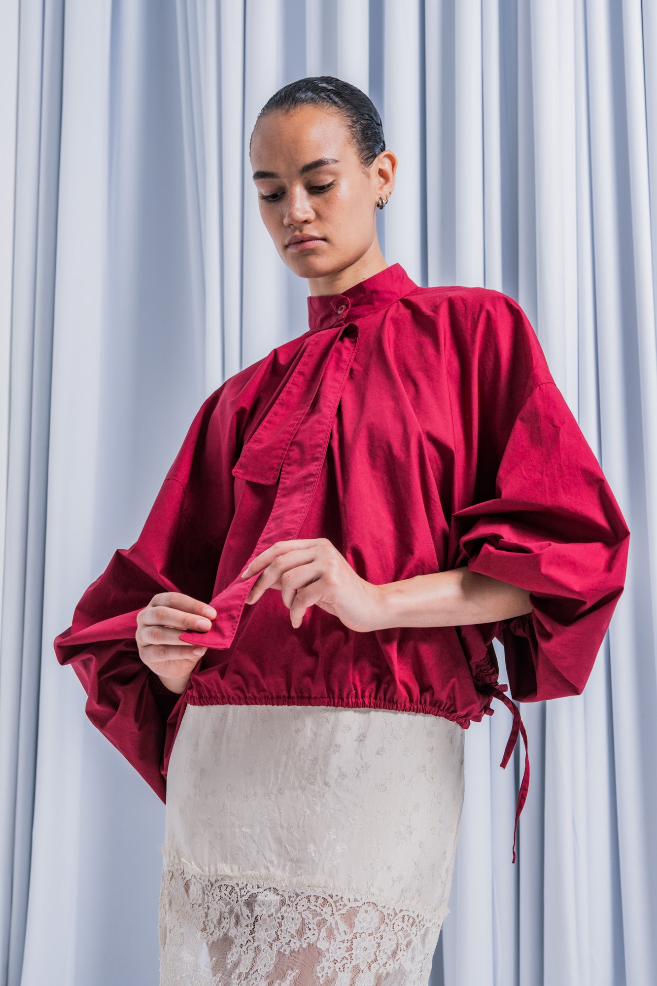 Woman wearing the Elva Blouse sewing pattern from Fibre Mood on The Fold Line. A top pattern made in poplin or silk fabric, featuring an oversized fit, stand-up collar, partial button placket with a detachable scarf, long sleeves, and elastic hem. 