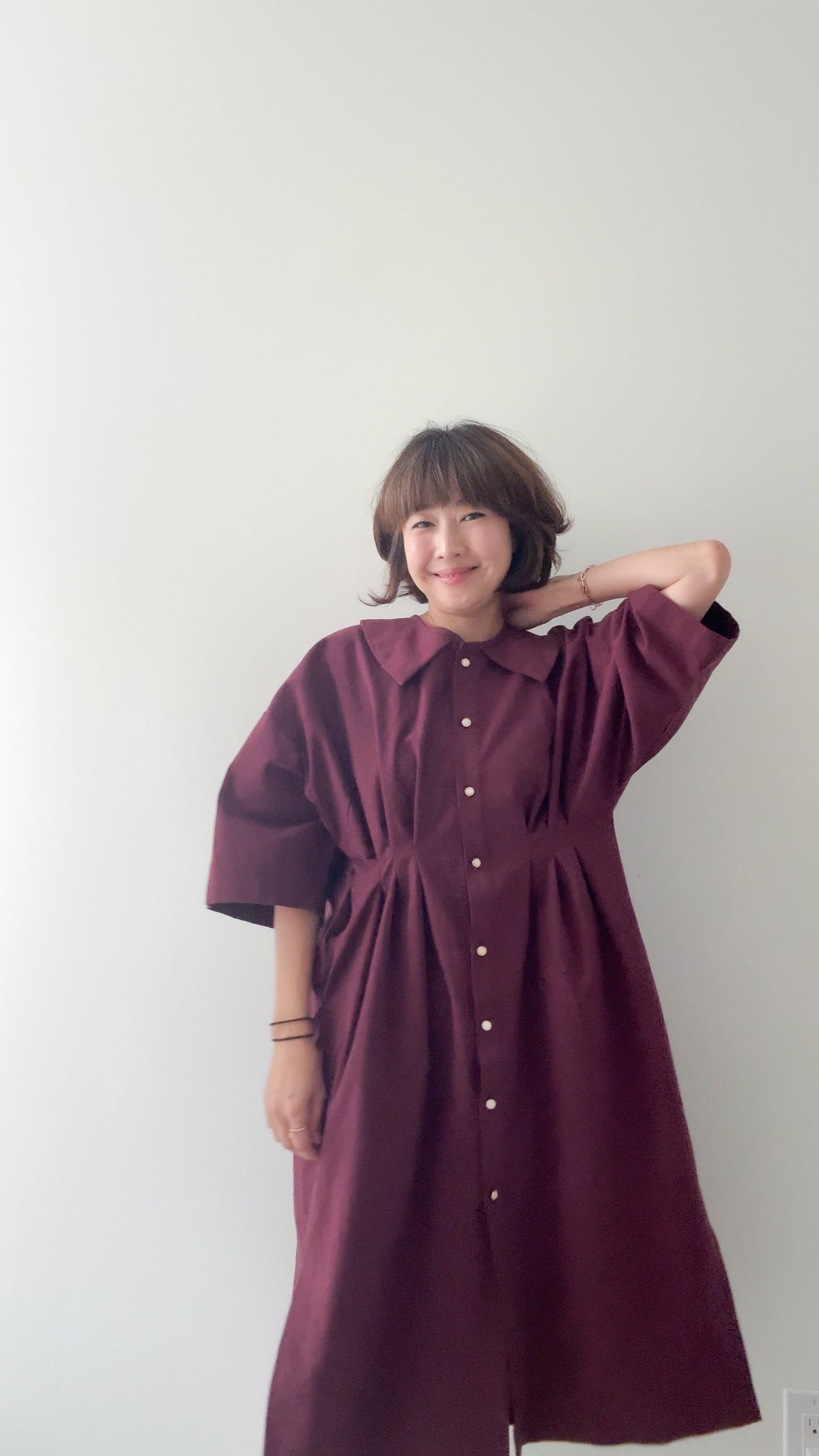 Woman wearing the Faux Collar Dress sewing pattern from Sewing Therapy on The Fold Line. A dress pattern made in cotton, linen or linen blend, tencel or rayon, wool or flannel, chambray or denim, or double gauze fabric, featuring an oversized fit, dropped shoulder, faux collar, button front, waist pleats, side slits, mid-length sleeves, and midi length.