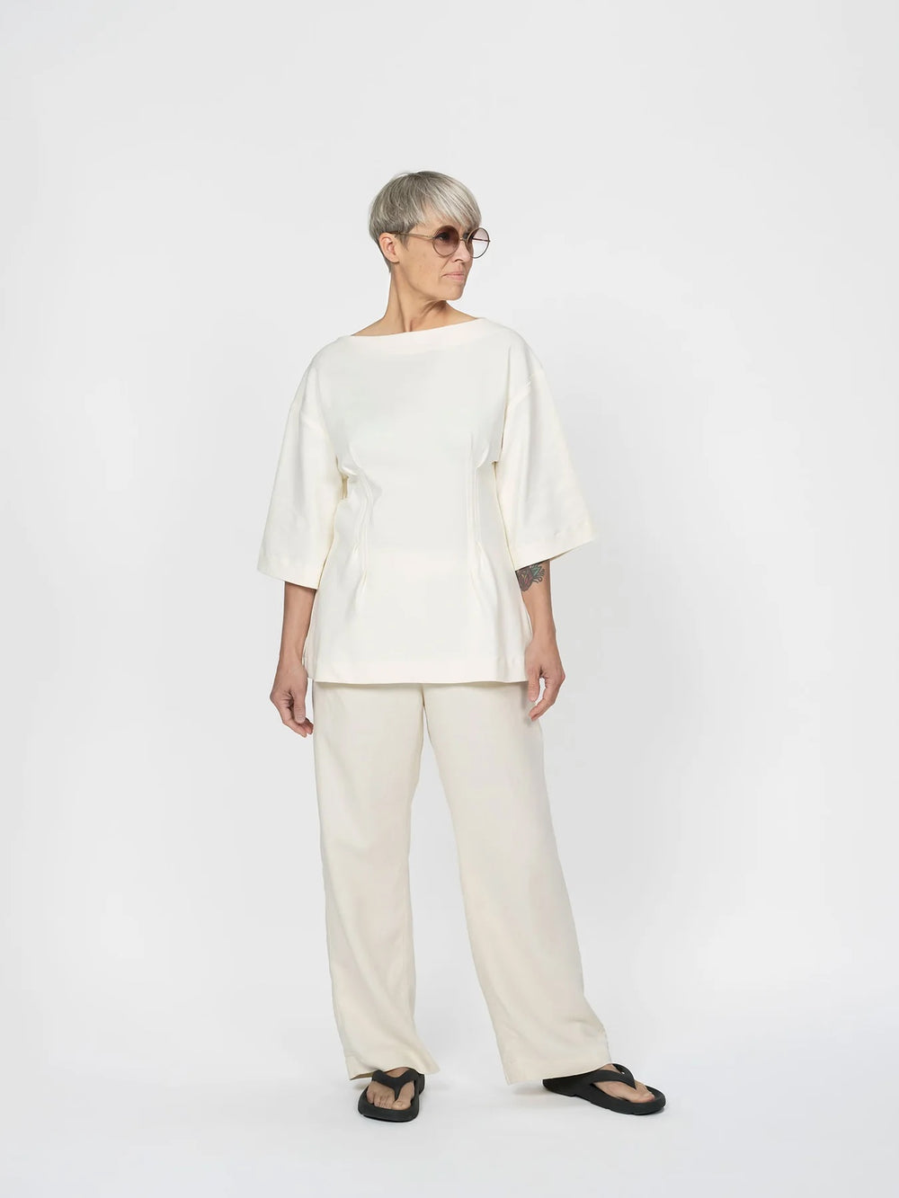 Woman wearing the Fitted Waist T-shirt sewing pattern from The Assembly Line on The Fold Line. A top pattern made in jersey fabric, featuring a boat neckline, dropped shoulders, wide elbow-length sleeves, and a shaped waist with topstitched pleats at the front and back.
