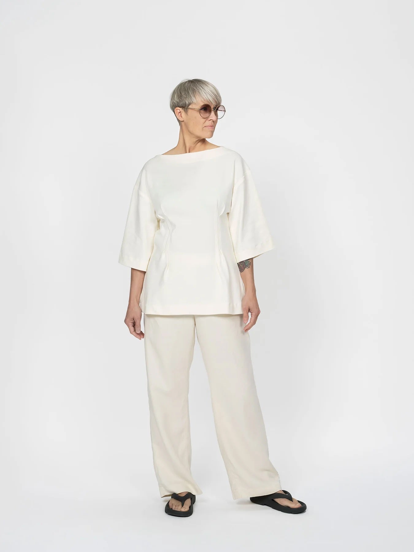 Woman wearing the Fitted Waist T-shirt sewing pattern from The Assembly Line on The Fold Line. A top pattern made in jersey fabric, featuring a boat neckline, dropped shoulders, wide elbow-length sleeves, and a shaped waist with topstitched pleats at the front and back.