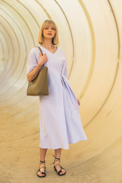 Woman wearing the Florrie Dress sewing pattern from Fibre Mood on The Fold Line. An asymmetric dress pattern made in viscose (crepe), chambray, crepon, satin, lyocell, seersucker, or cotton poplin or twill fabric, featuring a loose fit with a tie, V-neckline, wide short sleeves, and a midi length.