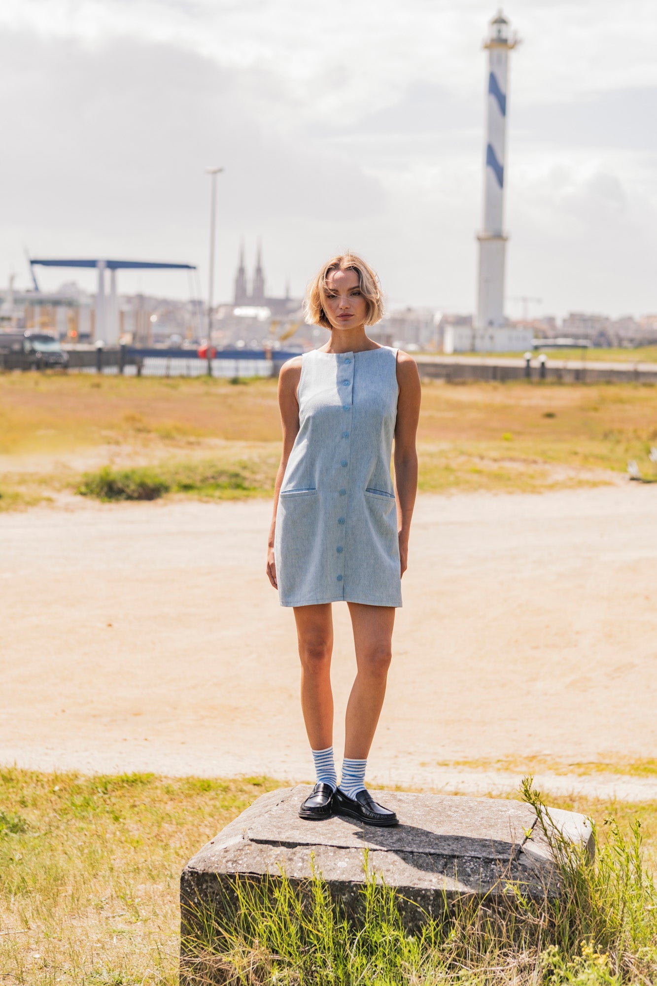 Woman wearing the Frankie Dress sewing pattern from Fibre Mood on The Fold Line. A sleeveless dress pattern made in cotton twill, denim, poly-cotton, fine corduroy, linen, or gabardine fabric, featuring a boat neck, button front and side zip, welt pockets, and mini length.