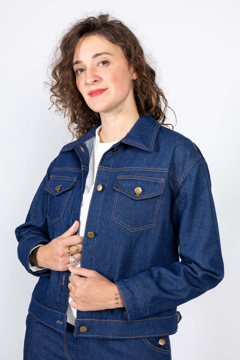 Woman wearing the Gold Jacket sewing pattern from I AM Patterns on The Fold Line. A jacket pattern made in denim, cotton canvas, gabardine, corduroy, cotton twill, or quilted fabric, featuring a straight cut, slightly dropped shoulders, shirt collar, front and back panelling, two flap chest pockets and two welt pockets, button closure, adjustable waist tabs, sleeves with cuffs, and contrast topstitching.