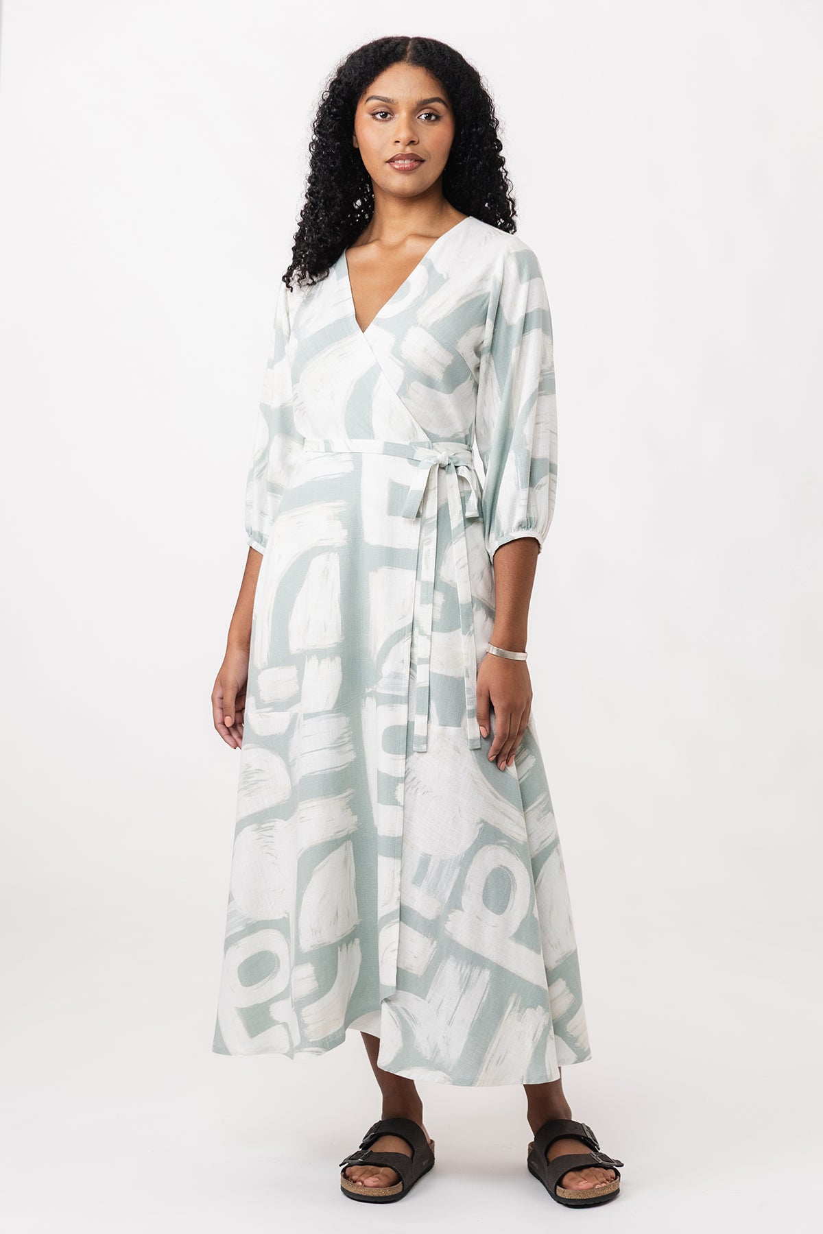Woman wearing the Hali Wrap Dress sewing pattern from Named on The Fold Line. A dress pattern made in cotton, linen, tencel, or rayon fabric, featuring a semi-fitted wrap bodice with a V-neck and back yoke, wrap tie closure, side pockets, 3/4 sleeves, and ankle length.