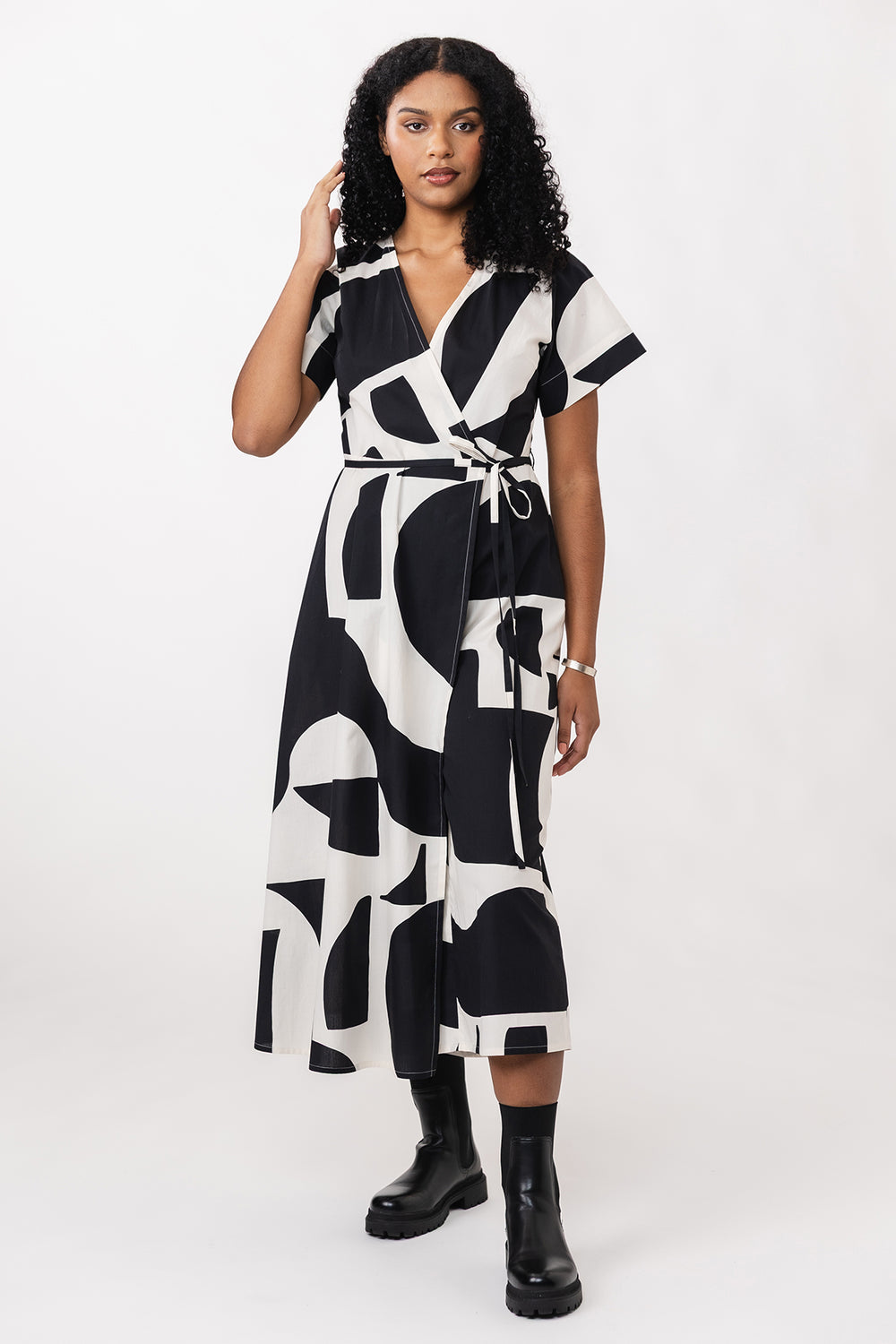 Woman wearing the Hali Wrap Jumpsuit sewing pattern from Named on The Fold Line. A jumpsuit pattern made in cotton, linen, tencel, or rayon fabric, featuring a semi-fitted wrap bodice with a V-neck, T-shirt sleeves, tie closure, side pockets, skirt panel, and midi length.