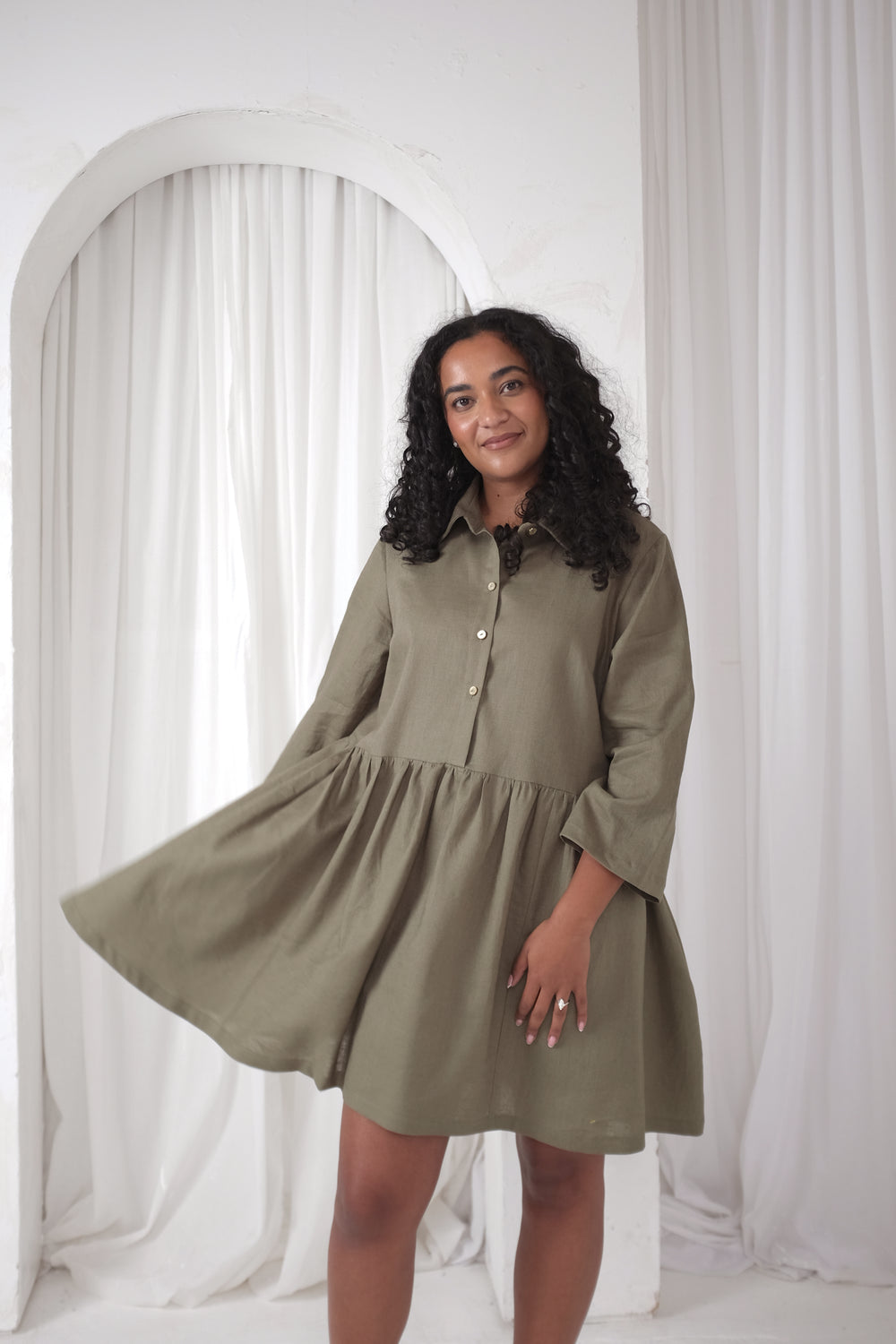 Woman wearing the Hampton Dress sewing pattern from NH Patterns on The Fold Line. A shirt dress pattern made in cotton, double gauze, linen and blends, or chambray fabric, featuring a loose fit, two-piece shirt collar, button front, 3/4 sleeves, gathered skirt, pockets, and above-the-knee length.