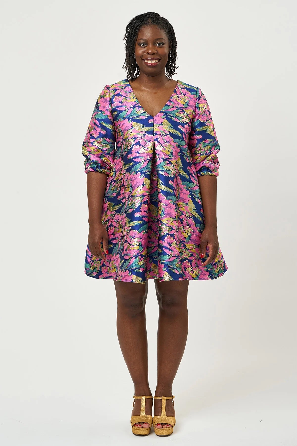 Woman wearing the Hettie Dress sewing pattern from Sew Over It on The Fold Line. A dress pattern made in brocade, jacquard, crepe, viscose, or cotton fabric, featuring a trapeze silhouette, V-neckline, pleat at the centre front, ties to cinch it in at the back, 3/4 sleeves, and a mini length. 