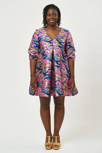 Woman wearing the Hettie Dress sewing pattern from Sew Over It on The Fold Line. A dress pattern made in brocade, jacquard, crepe, viscose, or cotton fabric, featuring a trapeze silhouette, V-neckline, pleat at the centre front, ties to cinch it in at the back, 3/4 sleeves, and a mini length. 