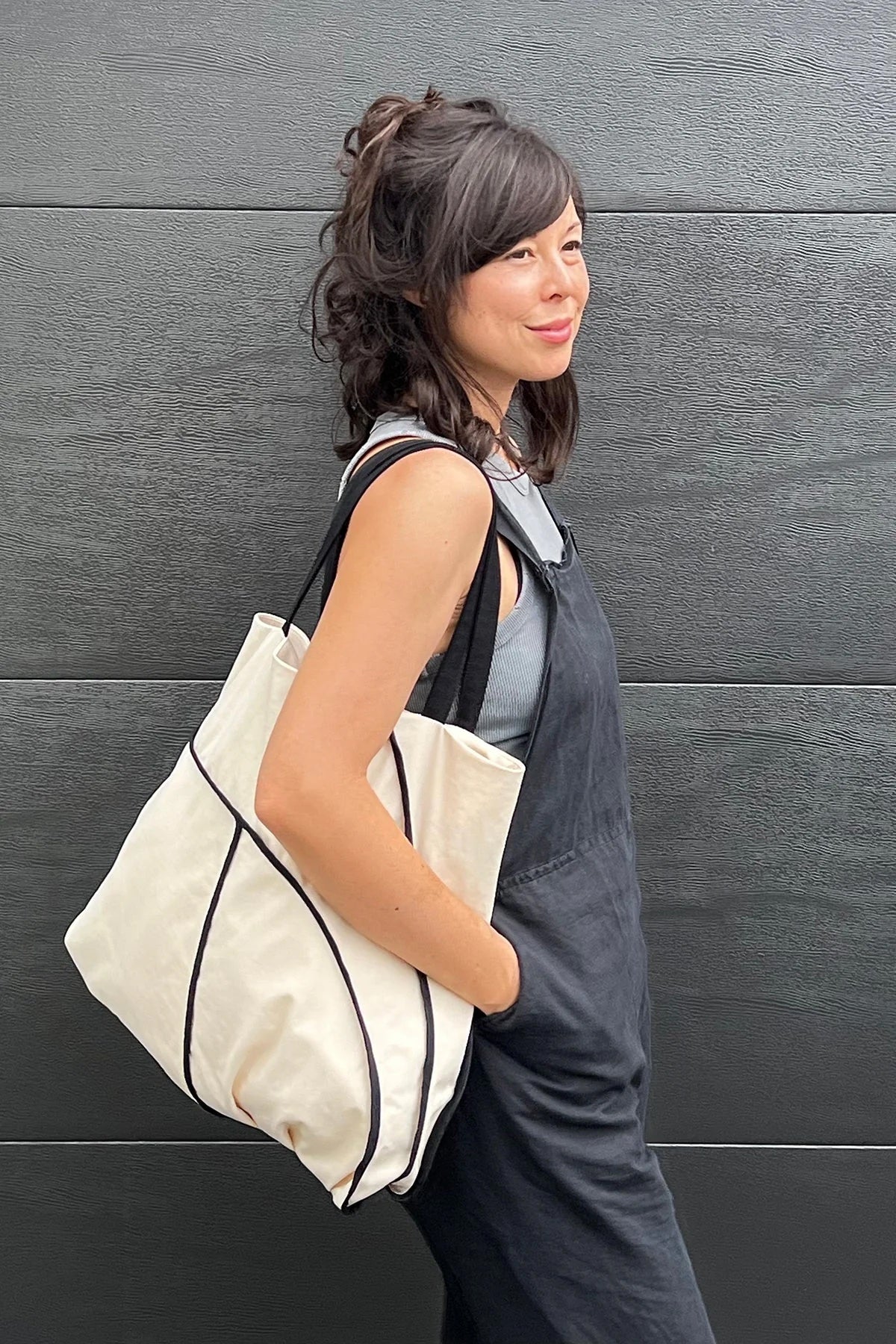 Woman carrying the Hove Tote sewing pattern from In the Folds on The Fold Line. A tote bag pattern made in linen, linen blend, cotton canvas, cotton drill, or denim fabric, featuring a puzzle-like panel construction, seam finishes on the outside, pleats, and a pocket.