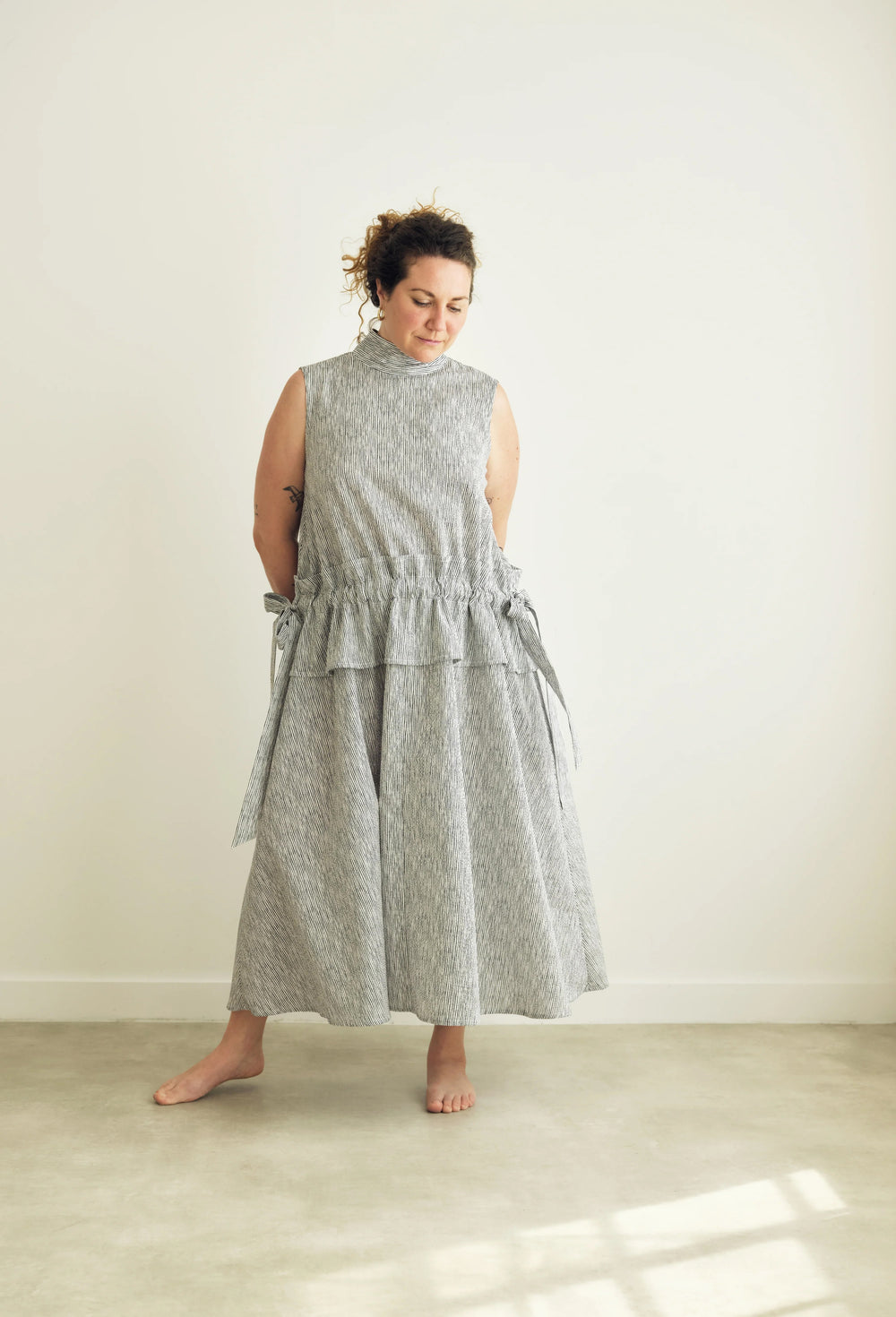 Woman wearing the Isabel Dress sewing pattern from the Modern Sewing Co. on The Fold Line. A sleeveless dress pattern made in cotton, seersucker, linen, viscose-linen/cotton blend, tencel, or silk fabric, featuring a high collar with ties, V-shaped back neckline, low waist, gathered waist panel with ties, ankle length skirt, and side pockets.