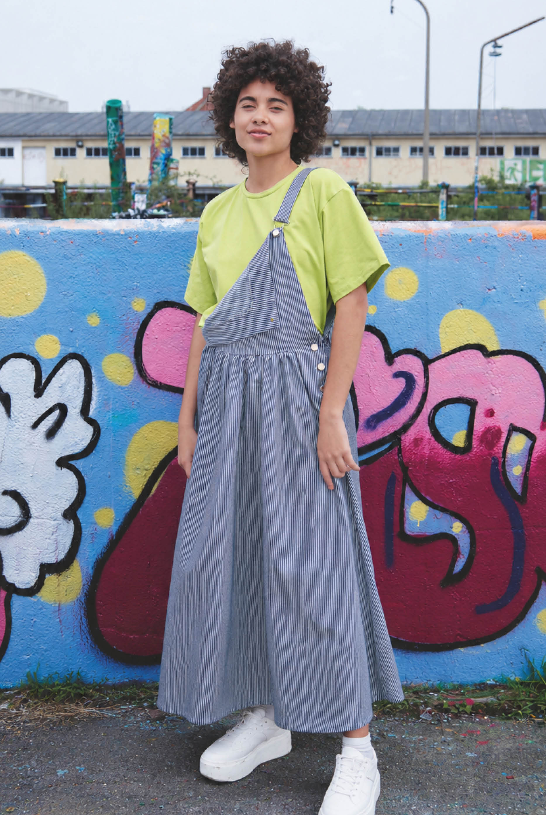 Woman wearing the Kylie Dungaree Dress sewing pattern from JULIANA MARTEJEVS on The Fold Line. A dress pattern made in linen, denim, or corduroy fabric, featuring a relaxed fit, bib with adjustable straps, gathered skirt, and midi length.