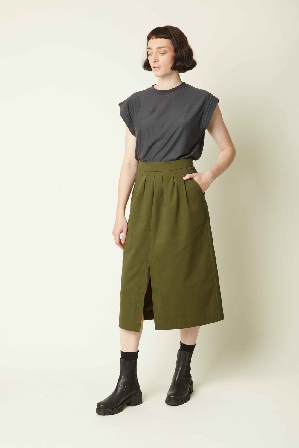 Woman wearing the La Jupe Défendue sewing pattern from Atelier Brunette on The Fold Line. A skirt pattern made in corduroy, gabardine, viscose, or viscose blend fabric, featuring a pleated front, front slit, side pockets, and midi length.