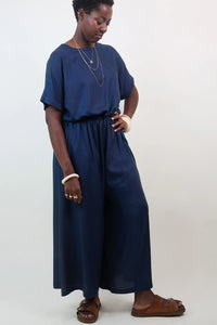 Woman wearing the La Robe Pantalon sewing pattern from Atelier Brunette on The Fold Line. A jumpsuit pattern made in viscose or viscose blend, linen, linen-viscose, cotton gauze, or lightweight cotton fabric, featuring a relaxed fit, round neckline, wide grown-on sleeves, back slit with a button, elastic waist, wide legs, side seam pockets, and 7/8 length. 