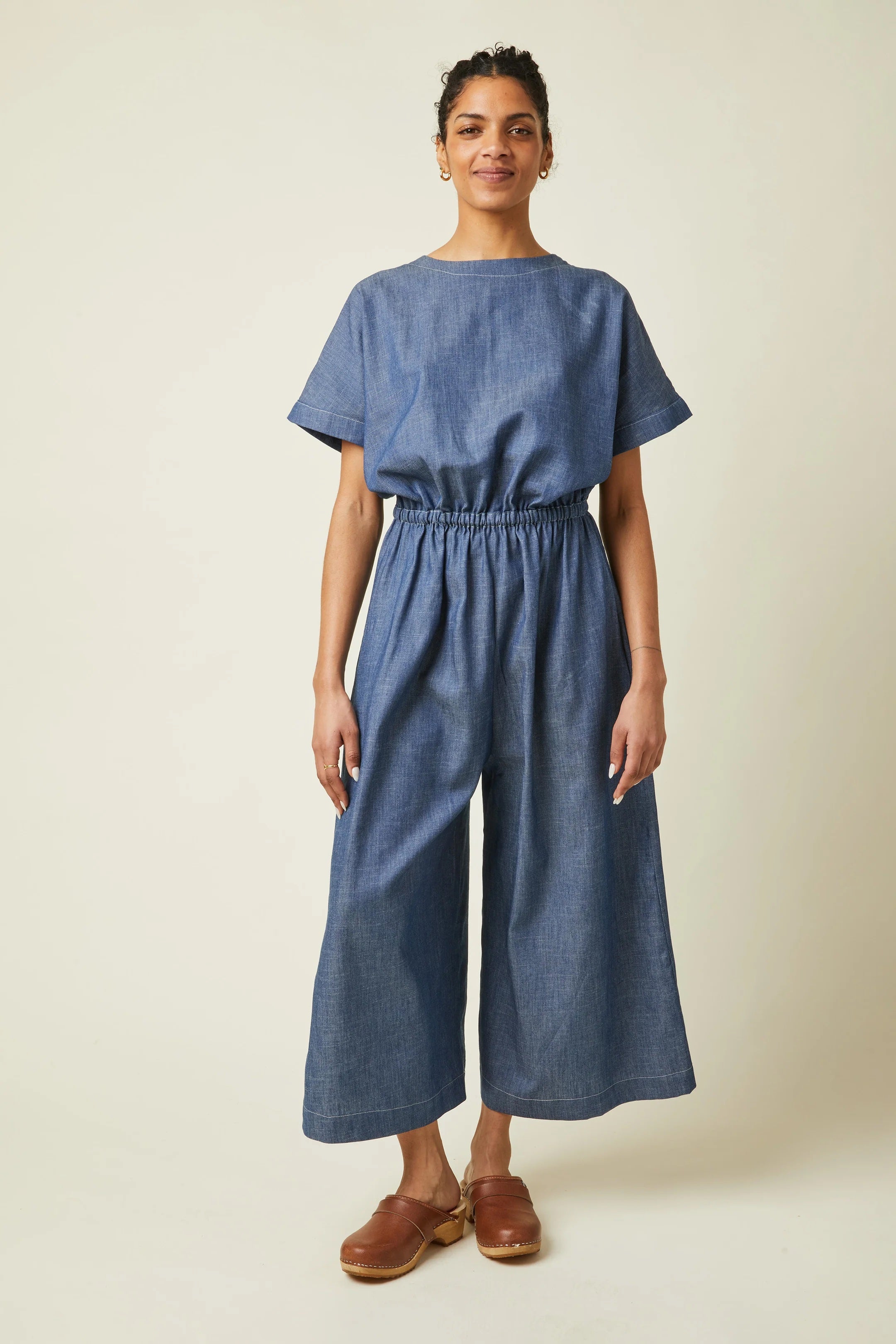Woman wearing the La Robe Pantalon sewing pattern from Atelier Brunette on The Fold Line. A jumpsuit pattern made in viscose or viscose blend, linen, linen-viscose, cotton gauze, or lightweight cotton fabric, featuring a relaxed fit, round neckline, wide grown-on sleeves, back slit with a button, elastic waist, wide legs, side seam pockets, and 7/8 length.