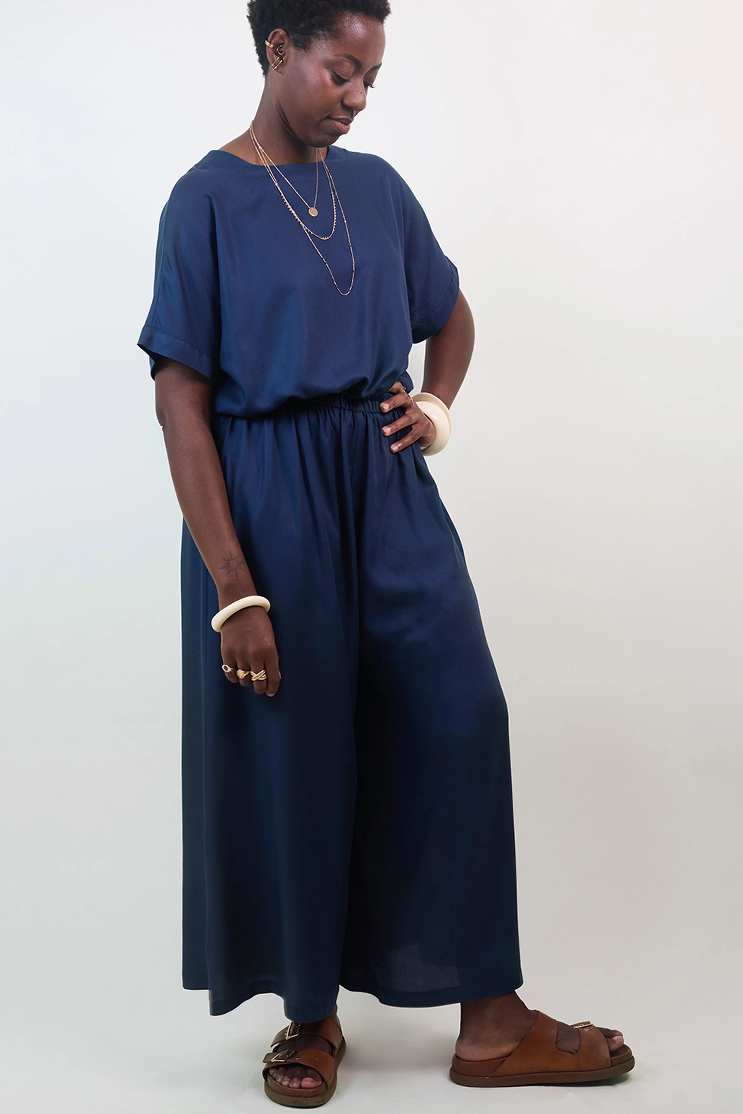 Woman wearing the La Robe Pantalon sewing pattern from Atelier Brunette on The Fold Line. A jumpsuit pattern made in viscose or viscose blend, linen, linen-viscose, cotton gauze, or lightweight cotton fabric, featuring a relaxed fit, round neckline, wide grown-on sleeves, back slit with a button, elastic waist, wide legs, side seam pockets, and 7/8 length. 