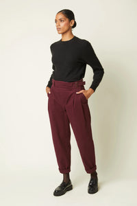 Woman wearing the Le Pantalon Ceinture sewing pattern from Atelier Brunette on The Fold Line. A trousers pattern made in corduroy or gabardine fabric, featuring a high waist, crossover closure, front pleats, side slant pockets, a back welt pocket, and tapered leg. 