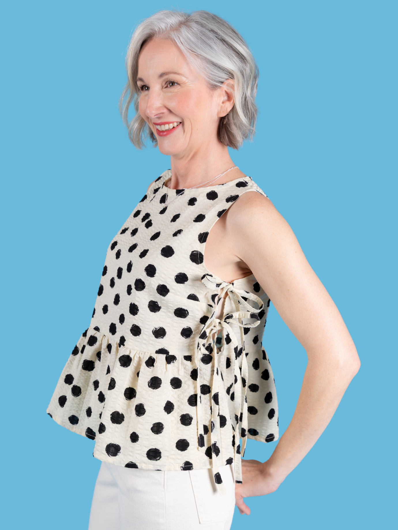 Woman wearing the Leah Top sewing pattern from Tilly and the Buttons on The Fold Line. A sleeveless top pattern made in cotton poplin, lawn, seersucker, double gauze, linen and blends, or viscose (rayon) fabric, featuring a round neckline, bust darts, side ties, and a gathered peplum.