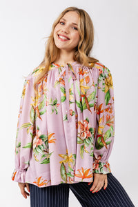 Woman wearing the Liia Gathered Blouse sewing pattern from Named on The Fold Line. A top pattern made in satin, viscose challis, or crepe fabric, featuring a relaxed fit, gathers along the yoke seam, front neckline ties, and long sleeves with ruffle cuffs.