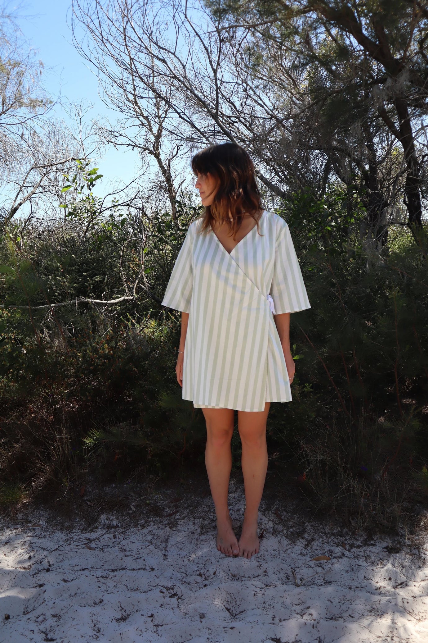 Woman wearing the Lilly Pilly Dress sewing pattern from Common Stitch on The Fold Line. A wrap dress pattern made in cotton or linen fabric, featuring a relaxed silhouette, V-neck, side tie, mid-length sleeves, and mini length.