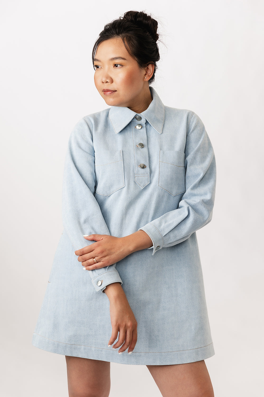 Woman wearing the Loviisa Denim Dress sewing pattern from Named on The Fold Line. A mini dress pattern made in denim or twill fabric, featuring an A-line silhouette, large shirt collar, chunky button placket, chest pockets, side pockets, long sleeves with cuffs, and topstitching.