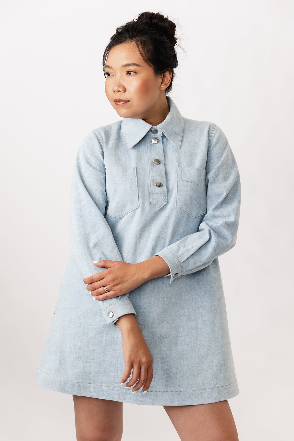 Woman wearing the Loviisa Denim Dress sewing pattern from Named on The Fold Line. A mini dress pattern made in denim or twill fabric, featuring an A-line silhouette, large shirt collar, chunky button placket, chest pockets, side pockets, long sleeves with cuffs, and topstitching.