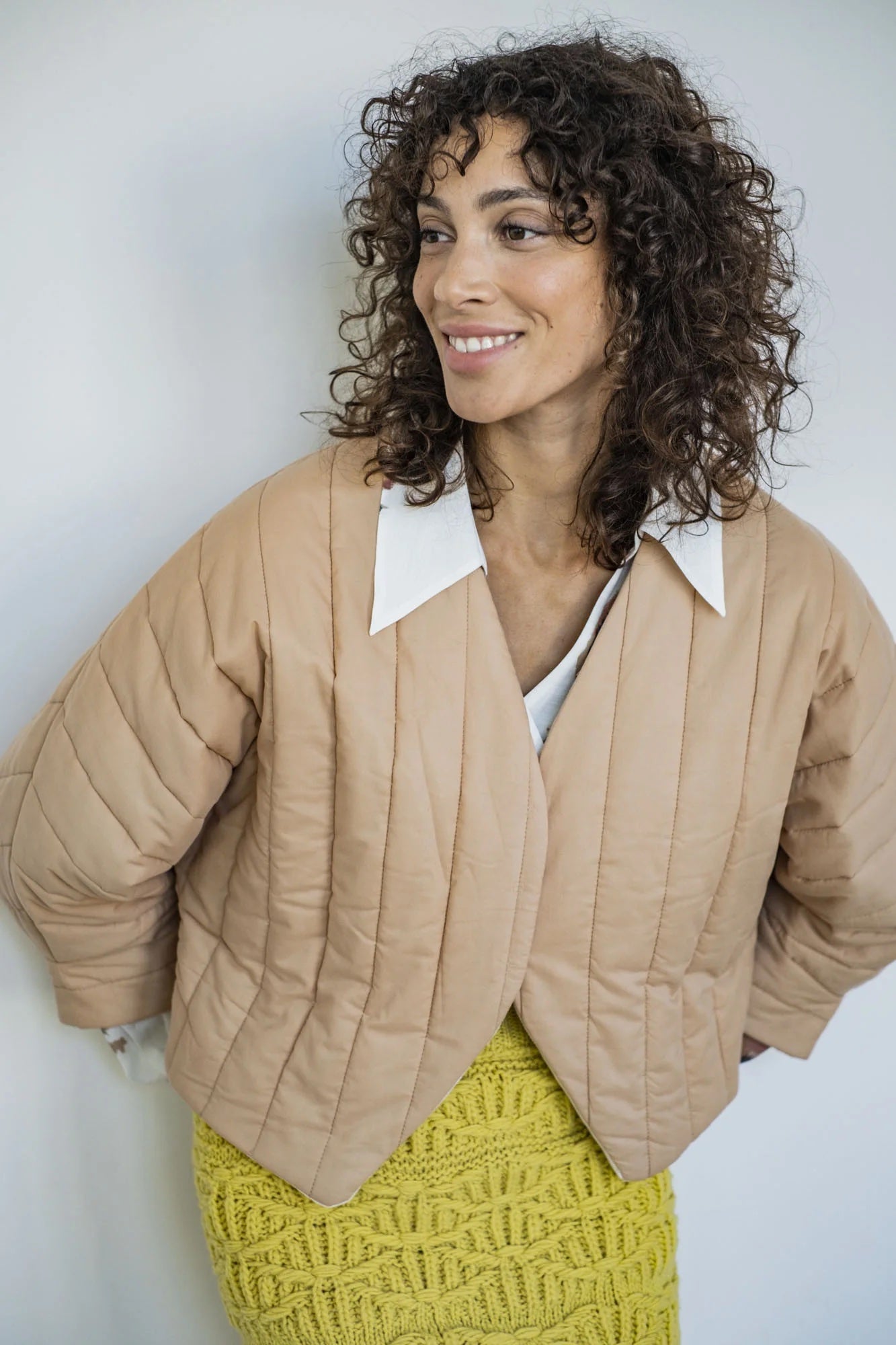Woman wearing the Lucy Jacket sewing pattern from Fibre Mood on The Fold Line. A jacket pattern made in quilted, knit, viscose crepe, lyocell, silk satin, jacquard, wool, corduroy, denim, leather, or suede fabric, featuring a V-neck, long sleeves with angled cuffs, hidden snap closure, and angled hem.
