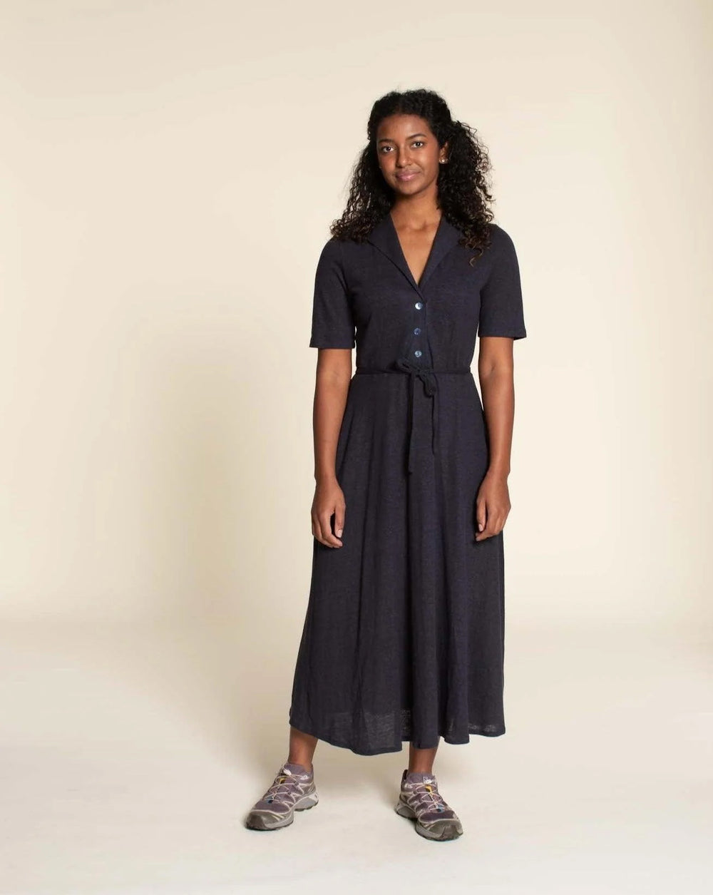 Woman wearing the Marianne Dress sewing pattern from Wardrobe by Me on The Fold Line. A dress pattern made in jersey fabric, featuring a fitted bodice with a shawl collar, button front, short sleeves, waist tie, and midi length. 